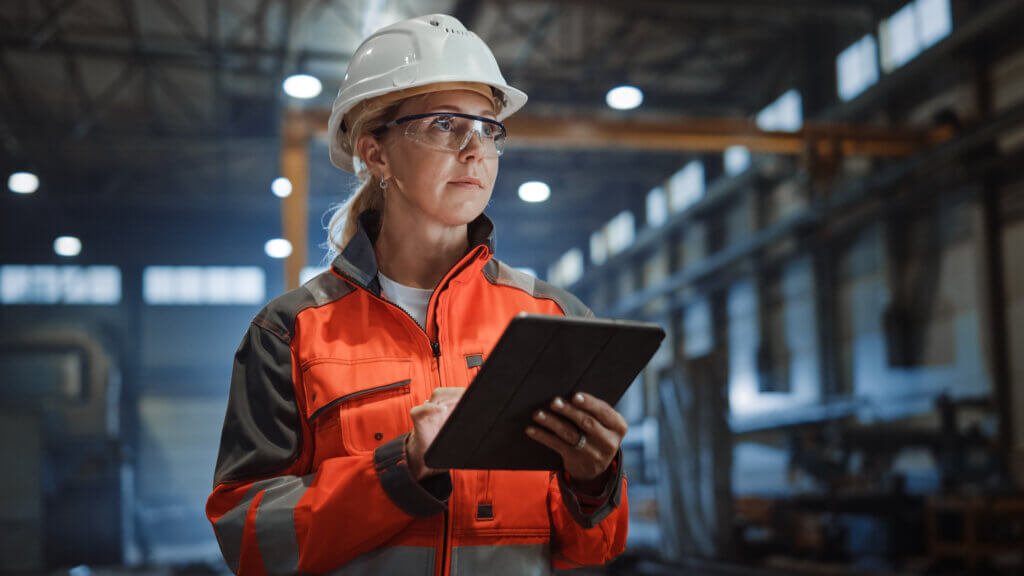 Ingenieurin mit Schutzhelm dokumentiert Angaben auf einem Tablet in einer Werkhalle